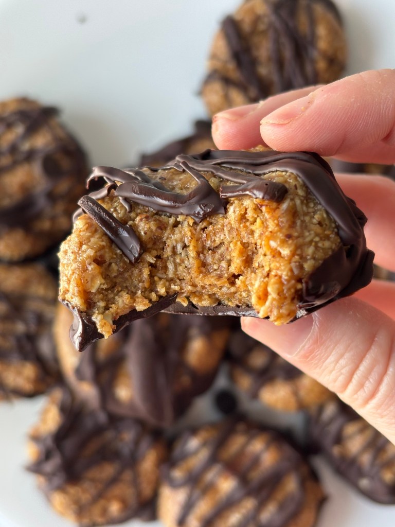 A close-up of a hand holding a chocolate-dipped cookie, showcasing a bite taken out to reveal its texture, with more cookies in the background.